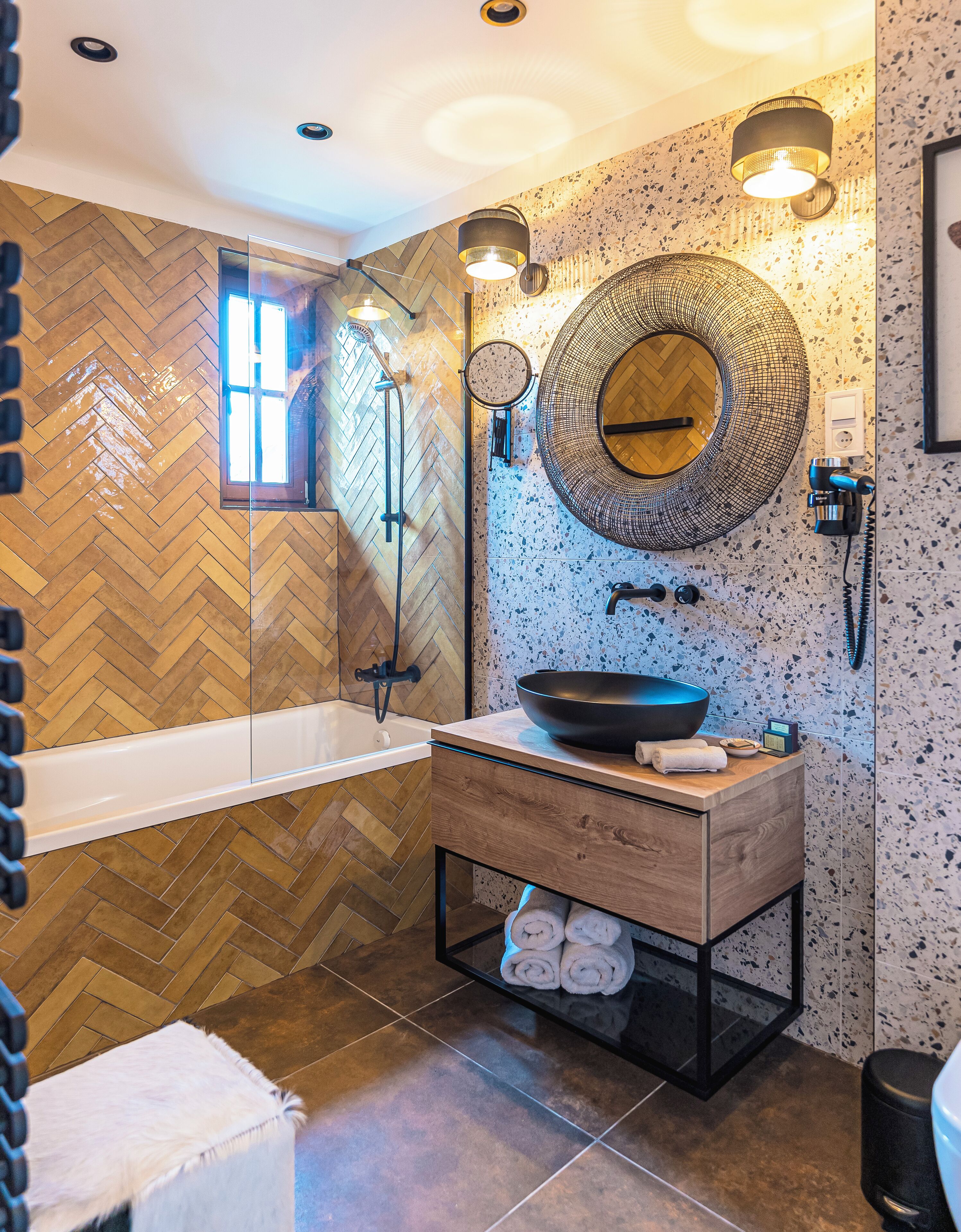 Un baño moderno con piso de madera en patrón de espiga, una bañera independiente, un espejo redondo y un lavamanos con lavabo negro.