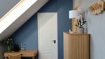 Una habitación acogedora con techo inclinado, una mesa de madera con sillas, una puerta blanca y un mueble pequeño con una lámpara y flores.
