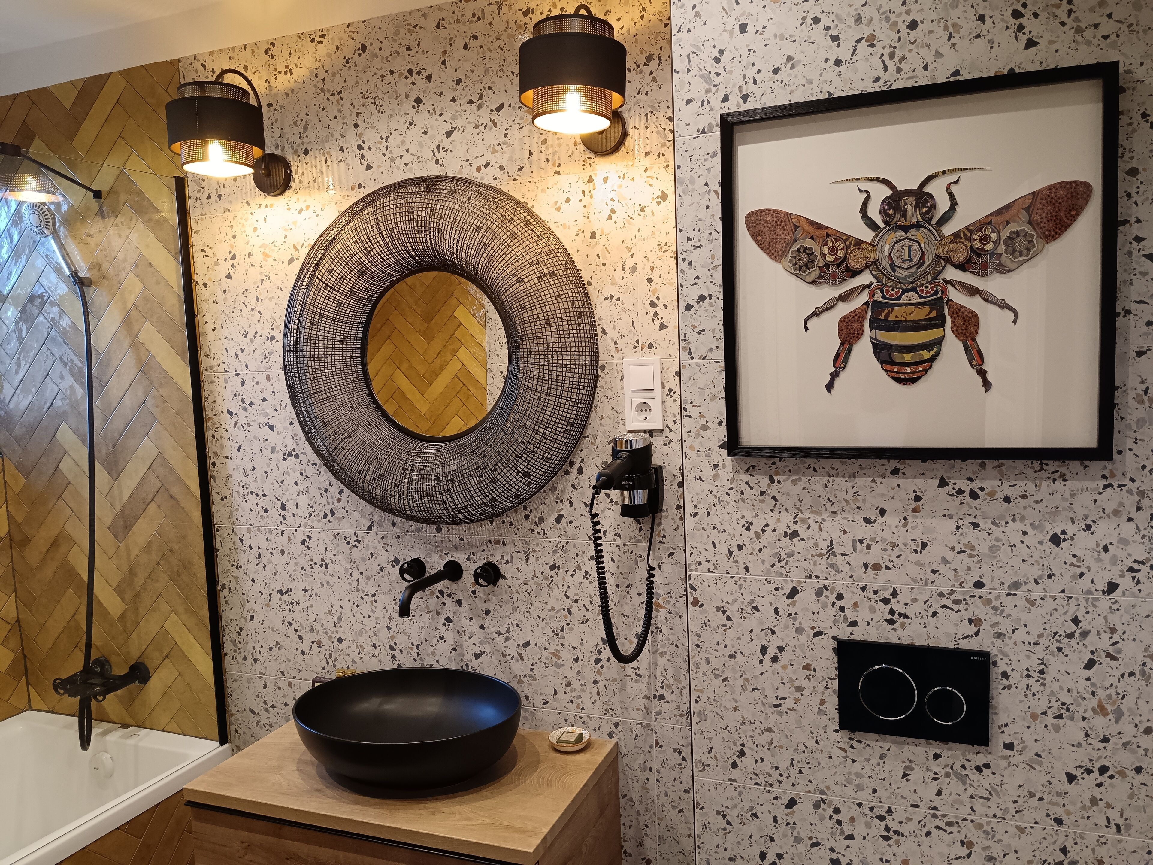 Une salle de bain avec un lavabo noir, un miroir à motifs en chevron, une œuvre d’art représentant une abeille encadrée et un porte-serviettes.