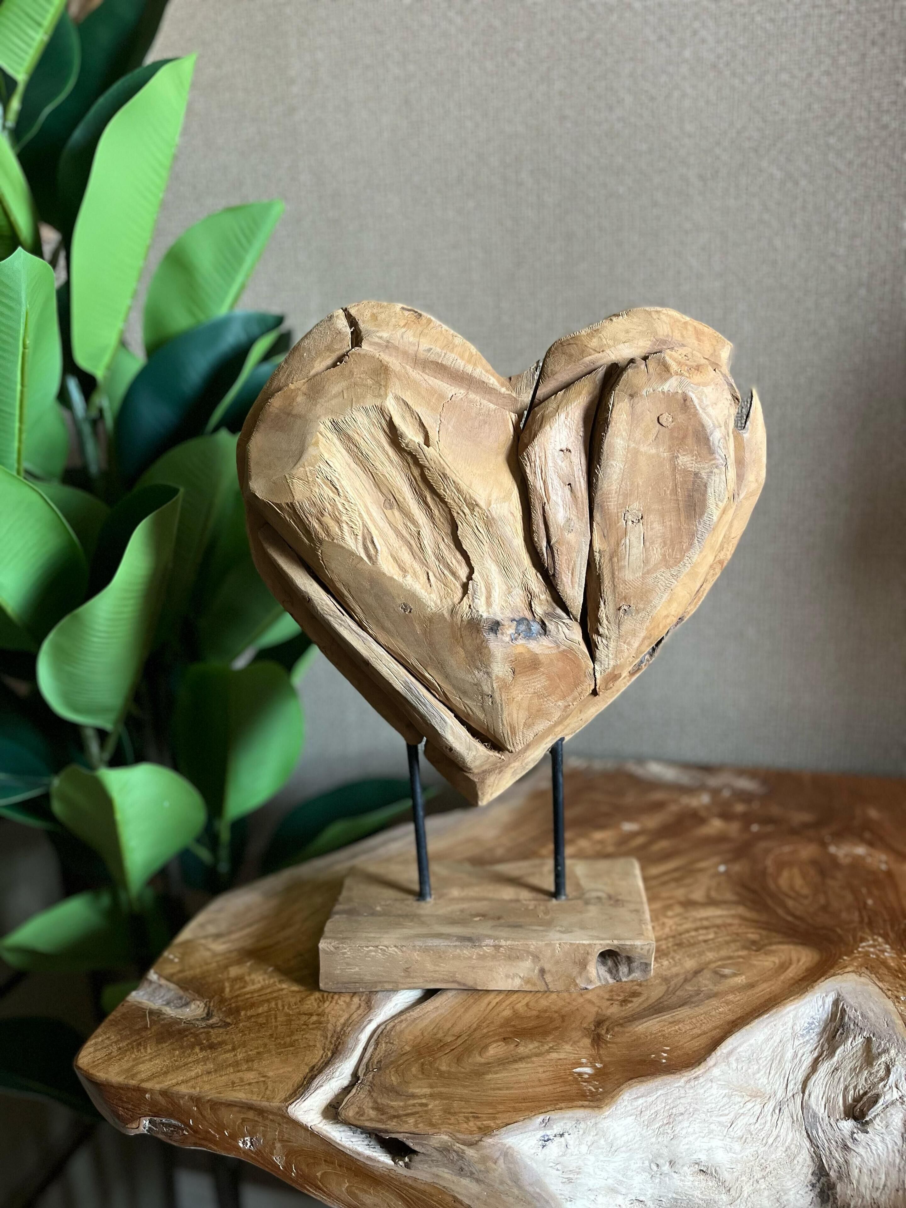Une sculpture en forme de cœur, faite de bois, est posée sur un socle, sur une surface également en bois, avec une plante en arrière-plan.