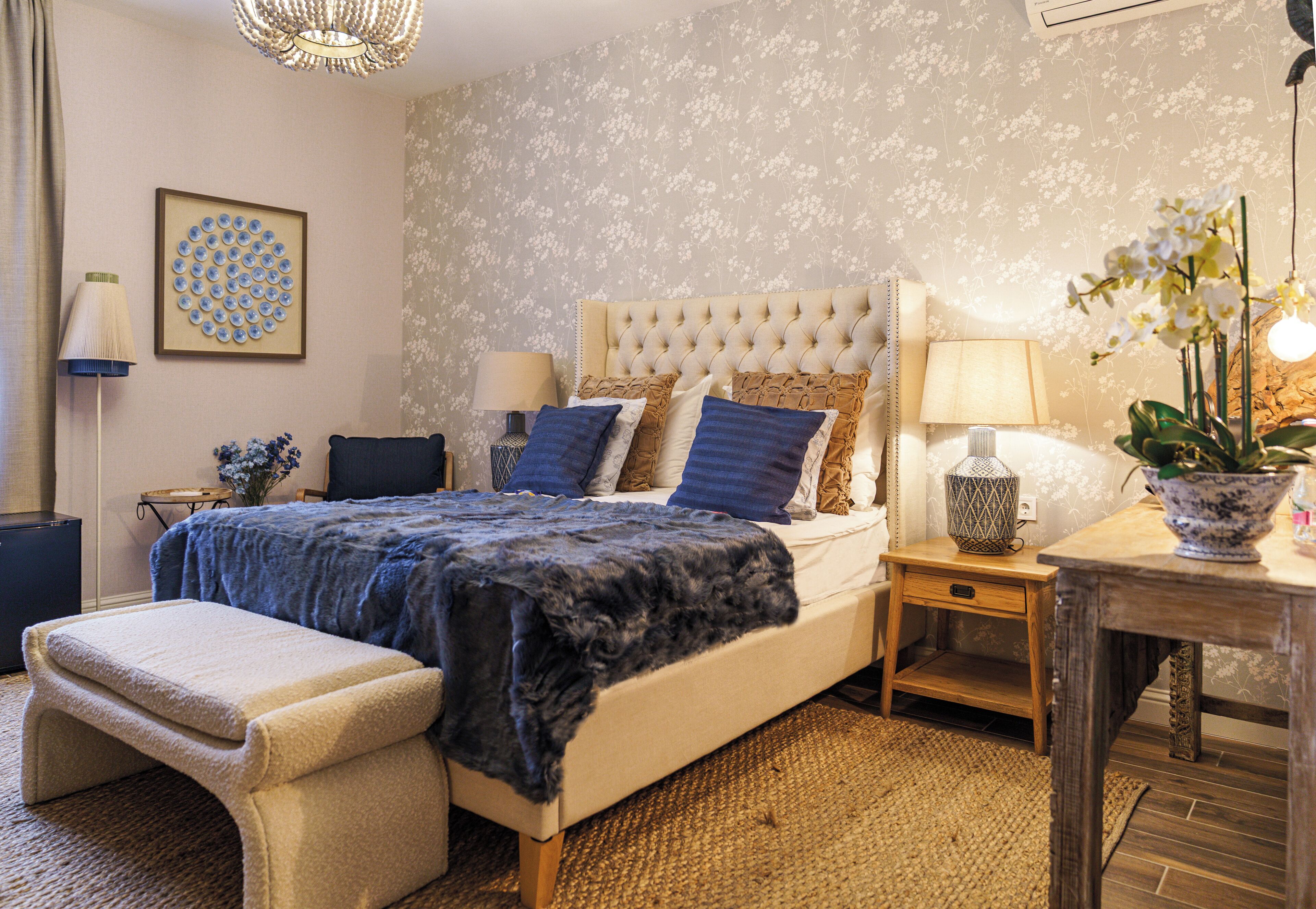 Een slaapkamer met een gestoffeerd hoofdeinde, een bed met blauwe en bruine beddengoed, een nachtkastje met een lamp, en een klein tafeltje met een plant.