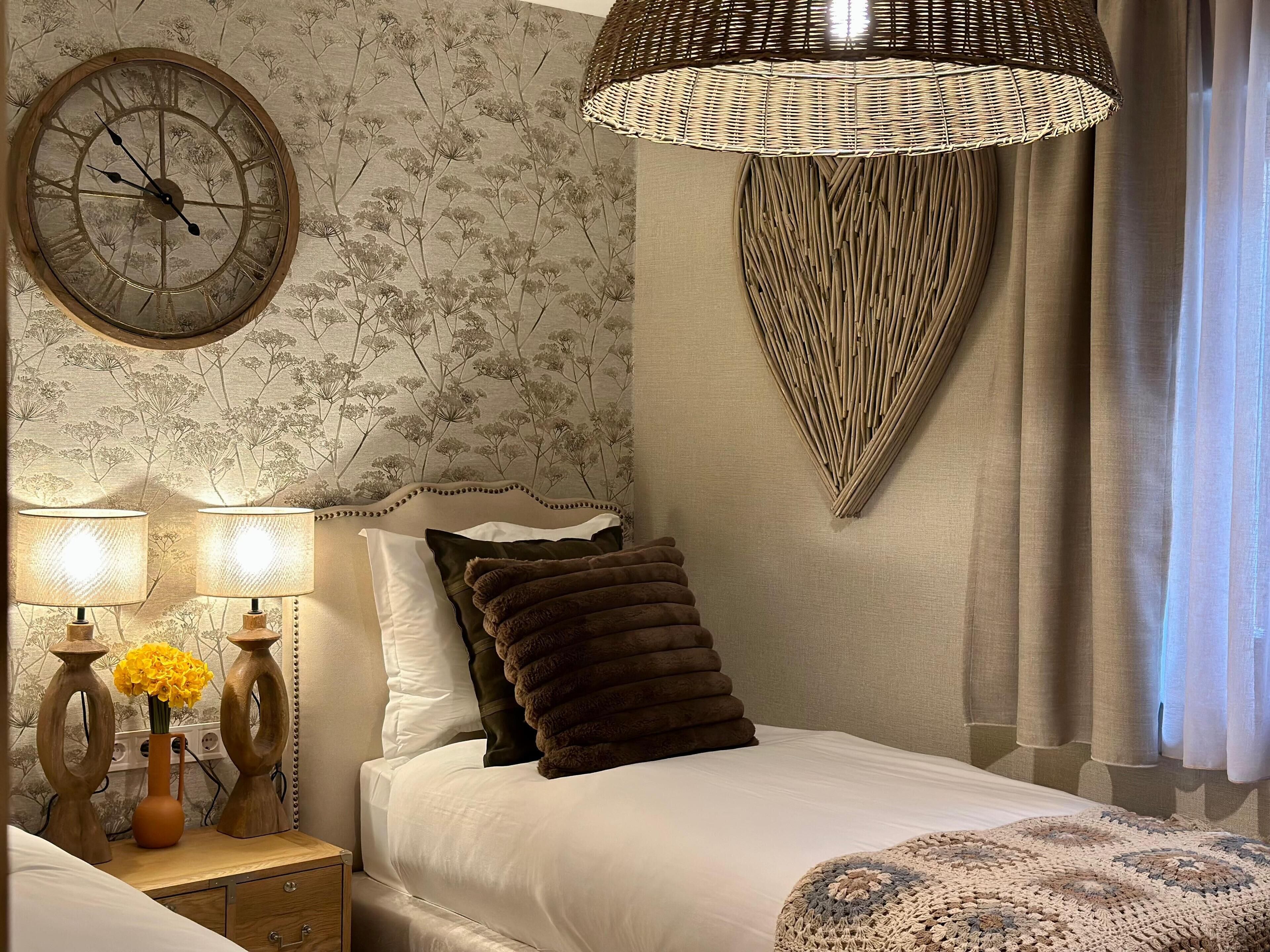 A bedroom with a large wall clock, a decorative wall hanging, a bedside lamp, and a bedside table with a vase of flowers.