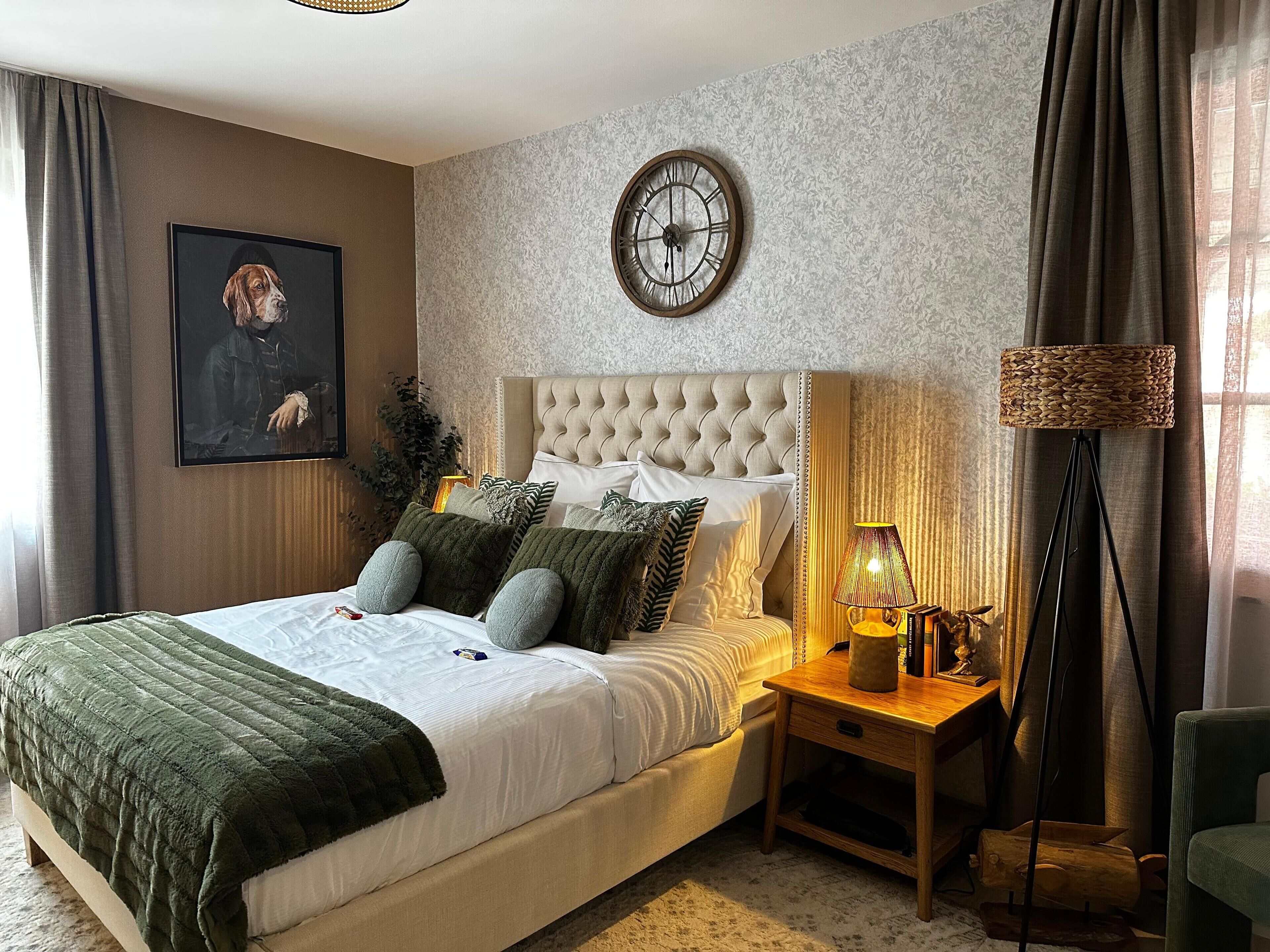 A neatly arranged bedroom with a large bed, a bedside lamp, a clock, and a framed picture on the wall.