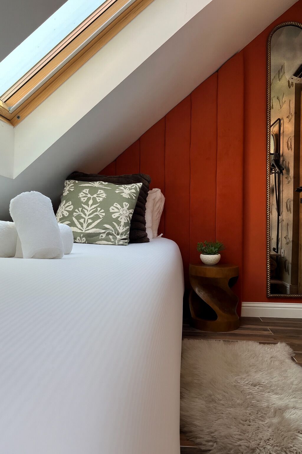 Een slaapkamer met een schuine plafond, een bed met wit beddengoed, een kussen met een groen en zwart patroon, een witte pluizige fauteuil, een klein rond tafeltje met een plant en een spiegel aan de muur.