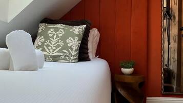Een slaapkamer met een schuine plafond, een bed met wit beddengoed, een kussen met een groen en zwart patroon, een witte pluizige fauteuil, een klein rond tafeltje met een plant en een spiegel aan de muur.