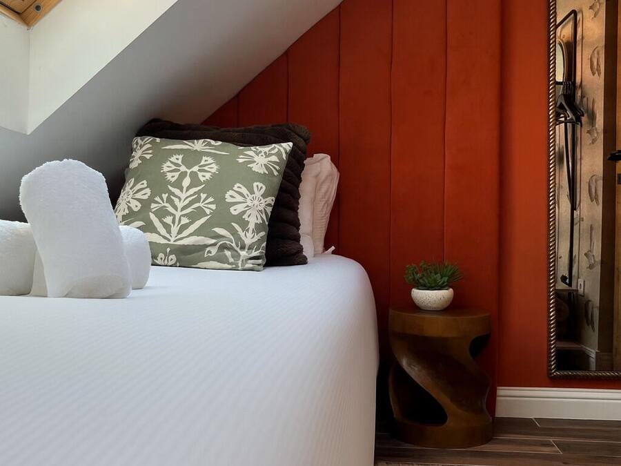A bedroom with a sloped ceiling, a bed with white bedding, a green and black patterned pillow, a white fluffy chair, a small round table with a plant, and a mirror on the wall.