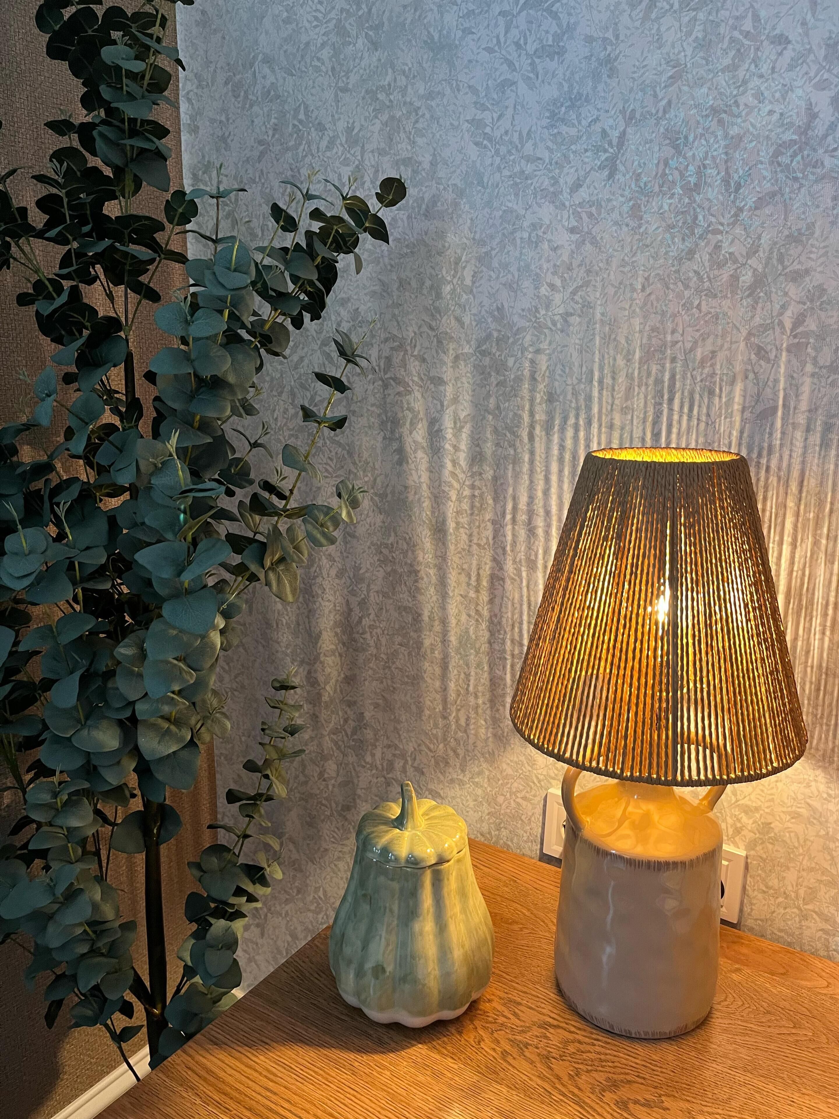 Une lampe avec un abat-jour à motifs en relief, posée sur une table en bois, à côté d’une plante aux grandes feuilles.