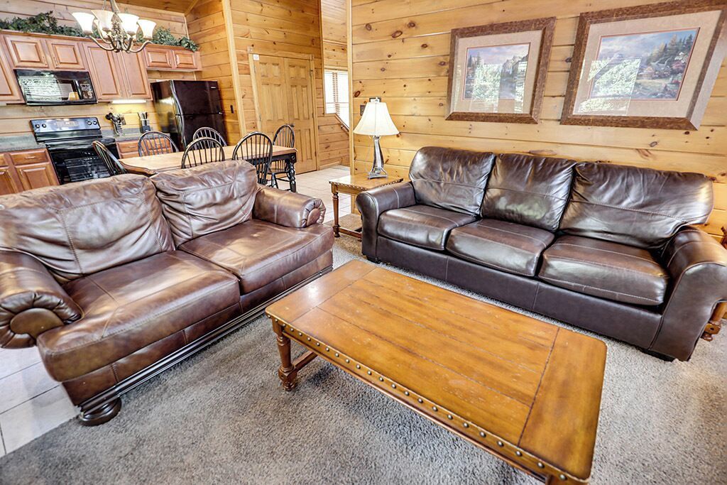 A log cabin interior with a brown leather sofa, a wooden coffee table, and a dining area with a table and chairs.