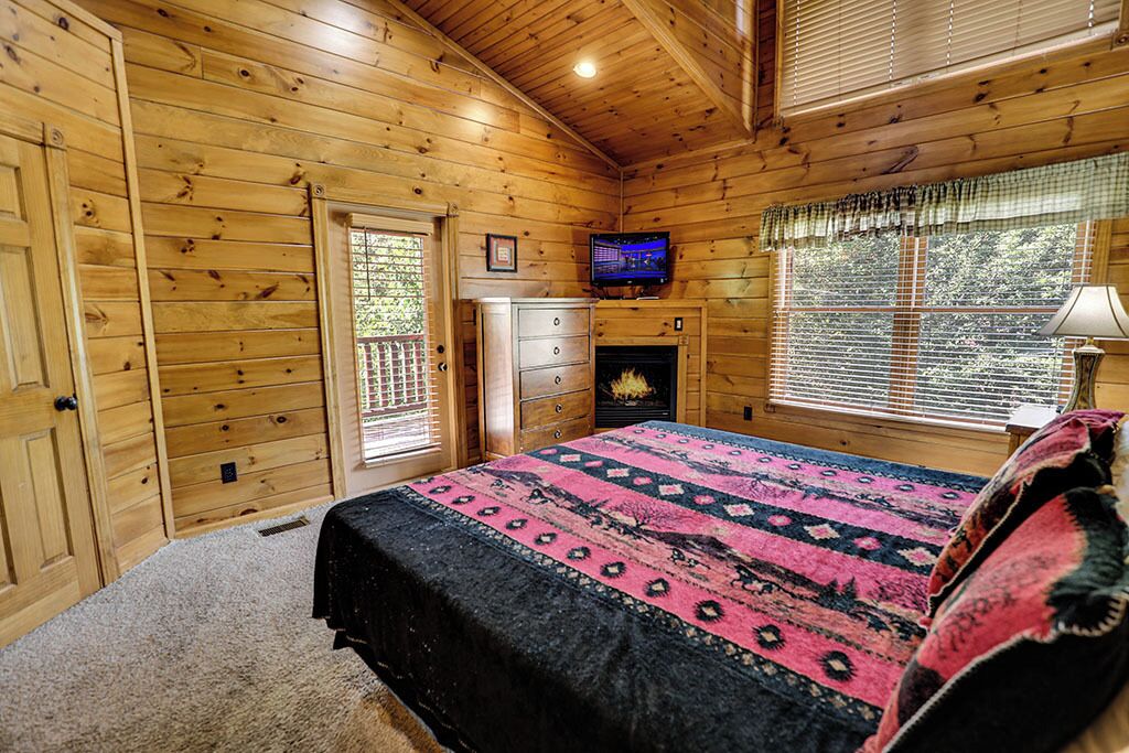 Ein Schlafzimmer in einer Blockhütte mit Bett, Fernseher, Kamin und einem Fenster mit Jalousien.