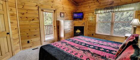 Ein Schlafzimmer in einer Blockhütte mit Bett, Fernseher, Kamin und einem Fenster mit Jalousien.