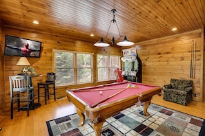 A wooden cabin interior with a pool table, chairs, a television, and a claw machine.