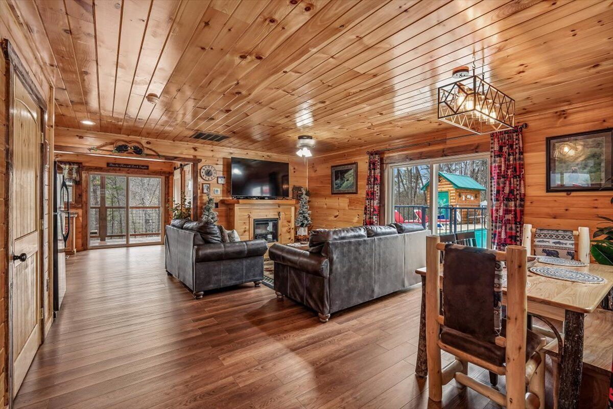 A log cabin interior with a fireplace, leather sofas, a dining table, and a TV.