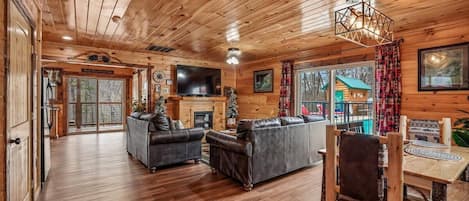 A log cabin interior with a fireplace, leather sofas, a dining table, and a TV.