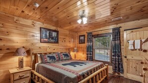 Quarto de cabana de madeira com cama, mesas de cabeceira, janela com cortinas e uma porta.