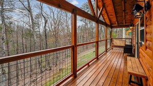 Um deck de madeira com guarda-corpo de metal, um banco e vista para uma floresta.