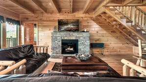 A cozy cabin interior with a stone fireplace, wooden staircase, and a leather sofa. - Smoky Mountain Charm w/ Hot Tub & Views (Sevierville)