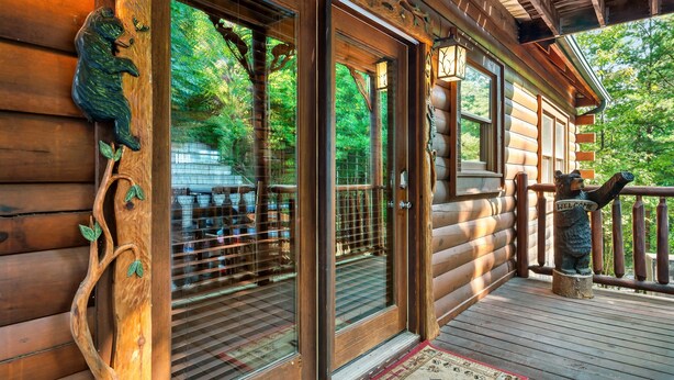 A wooden cabin with a glass door, a wooden deck, and a bear statue on the railing. - Smoky Mountain Charm w/ Hot Tub & Views (Sevierville)