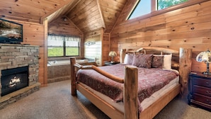 A log cabin bedroom with a stone fireplace, a large bed, a wooden ceiling, and a view of the outdoors. - Smoky Mountain Charm w/ Hot Tub & Views (Sevierville)