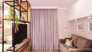 Uma sala de estar moderna com um sofá, uma televisão sobre um suporte de madeira, uma janela grande com cortinas e uma estante com plantas.