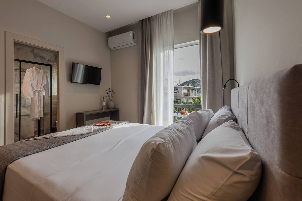 A hotel room with a large bed, a flat-screen TV, an air conditioning unit, a window with curtains, and a view of buildings outside.