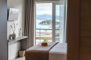 A hotel room with a bed, a TV mounted on the wall, a view of a body of water and mountains, and a balcony with a railing. - Noma Suites Tolo (Tolo)