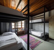 A dormitory room with bunk beds, a wooden ceiling, a window with curtains, a colorful rug, and a small desk with a lamp.