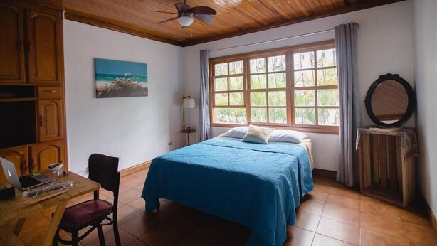 A bedroom with a wooden ceiling, a bed with a blue bedspread, a desk with a laptop, and a painting on the wall. - The Garden CR (San Pedro)