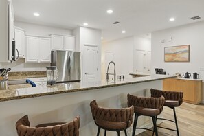 A modern kitchen with white cabinets, a stainless steel refrigerator, a granite countertop, and a bar area with brown upholstered stools.