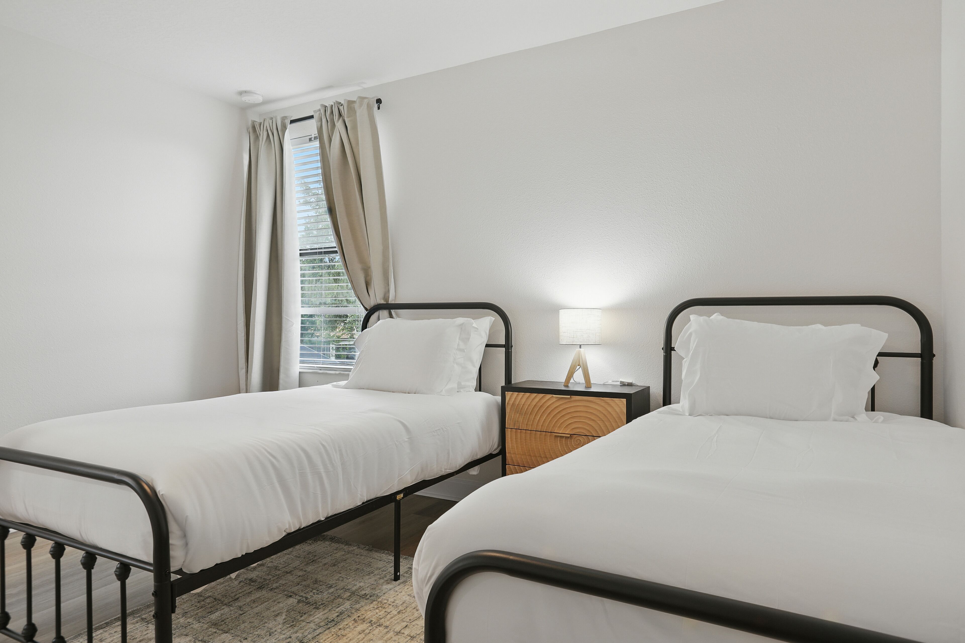 A room with two single beds, a nightstand with a lamp, and a window with curtains.