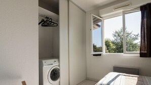 A compact room with a washing machine, a bed with striped bedding, and a window with a view of trees.