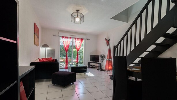 A modern living room with a sofa, ottoman, and a TV. There is a staircase to the right and a window with curtains.