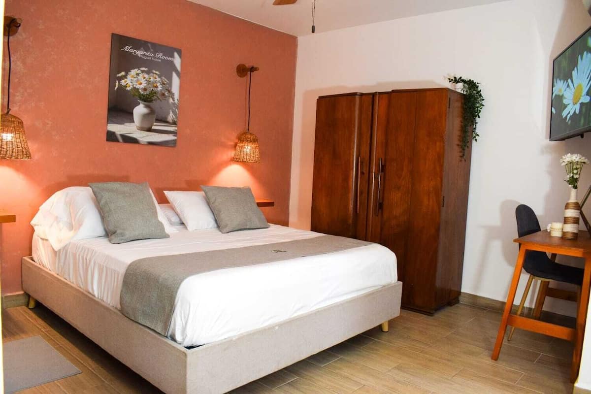 A bedroom with a bed, a wooden wardrobe, a desk with a chair, and a poster on the wall.