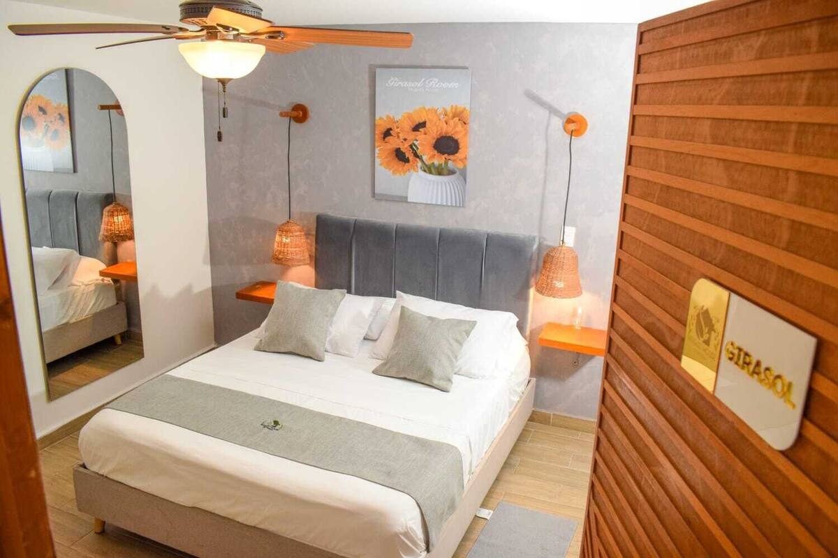 A bedroom with a bed, a ceiling fan, a wall-mounted shelf with a vase of sunflowers, and a wooden panel wall with a sign.