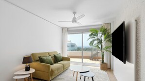 Ein modernes Wohnzimmer mit einer Couch, einem kleinen Tisch, einer Lampe und einem großen Fenster mit Blick auf Gebäude und eine Palmenbaum.