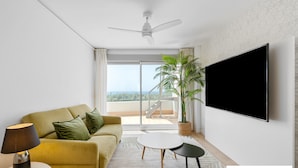 Ein modernes Wohnzimmer mit einer Couch, einem Fernseher, einem Deckenventilator und Blick ins Freie.