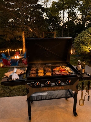 Una parrilla Blackstone con un pozo de fuego, mobiliario exterior y vistas al jardín trasero.