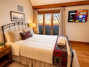 Una cama bien hecha con colcha y almohadas, una mesita de noche con lámpara, un televisor de pared que muestra servicios de streaming y una ventana con cortinas.