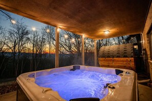 Un jacuzzi bajo una pérgola de madera con luces colgantes, rodeado de árboles y una estructura de madera.