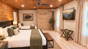 A bedroom with a large bed, a smaller bed, a bench, a ceiling fan, a brick wall, a painting, and a window with curtains.