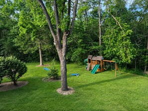 Un patio trasero con una casita de juegos de madera, un columpio y un árbol pequeño.