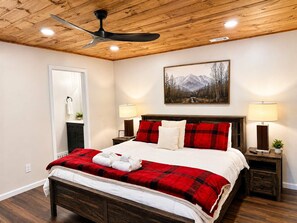 Un dormitorio con techo de madera, cama con ropa de cama roja y blanca, dos lámparas de noche, un cuadro de montañas y piso de madera.