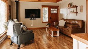 A living room with a wooden ceiling, a television mounted on the wall, a brown leather sofa, a grey armchair, and a wooden coffee table.