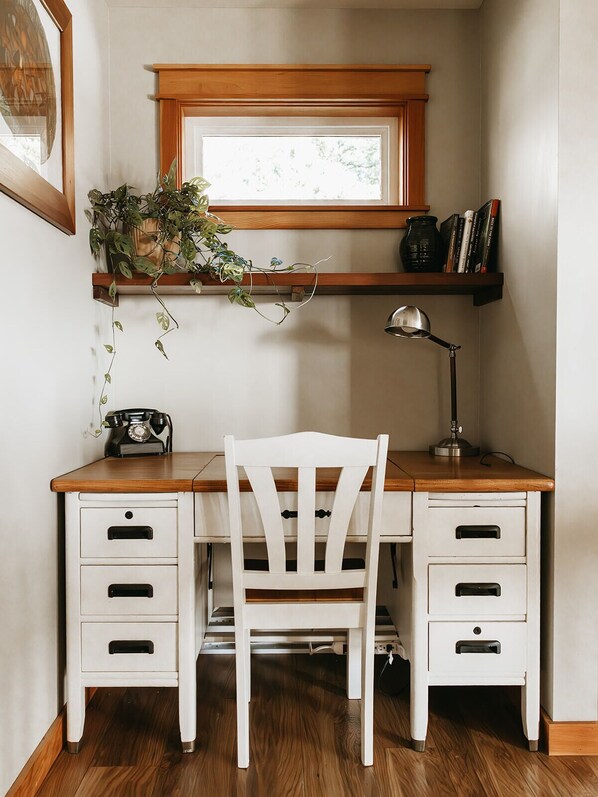 Un estudio con escritorio de madera, cajones blancos, una silla blanca, teléfono antiguo, lámpara de escritorio, ventana con marco de madera, estantería con libros y planta.