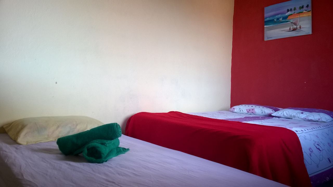 A single bed with a red blanket and a white sheet, a green towel, and a beige pillow.