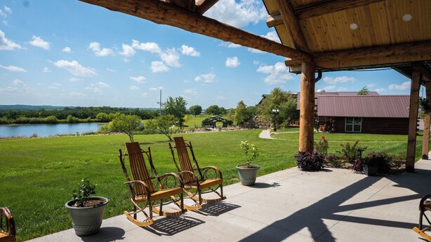 A covered patio with wooden chairs and potted plants, overlooking a grassy area and a body of water. - Our beautiful lodge near Albany, MO is sure to please! Perfect for large groups (Albany)