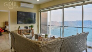 A living room with a large TV, wicker furniture, and a view of a body of water and mountains. - Stunning Lake-view Villa at Coatepeque - Wifi, A/C (El Congo)