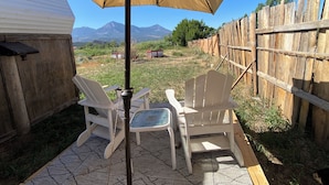 Un patio con dos sillas blancas y una mesa bajo un paraguas color beige, una cerca de madera y una caravana al fondo.