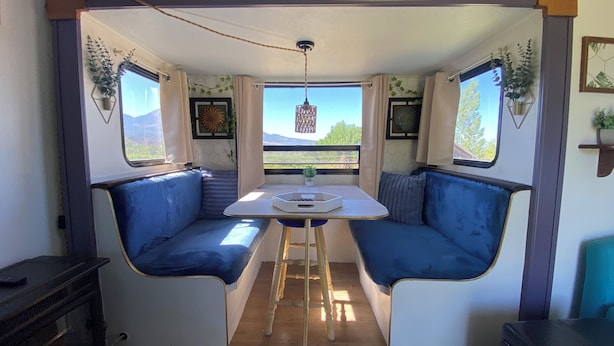 Interior acogedor de una autocaravana con dos zonas de asientos tapizadas en azul alrededor de una mesa central, una escalera pequeña y una ventana con vistas a vegetación y montañas.