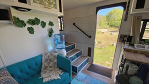 Un espacio habitable compacto con un sofá azul, un dispensador de agua y vistas a una zona verde a través de una puerta.