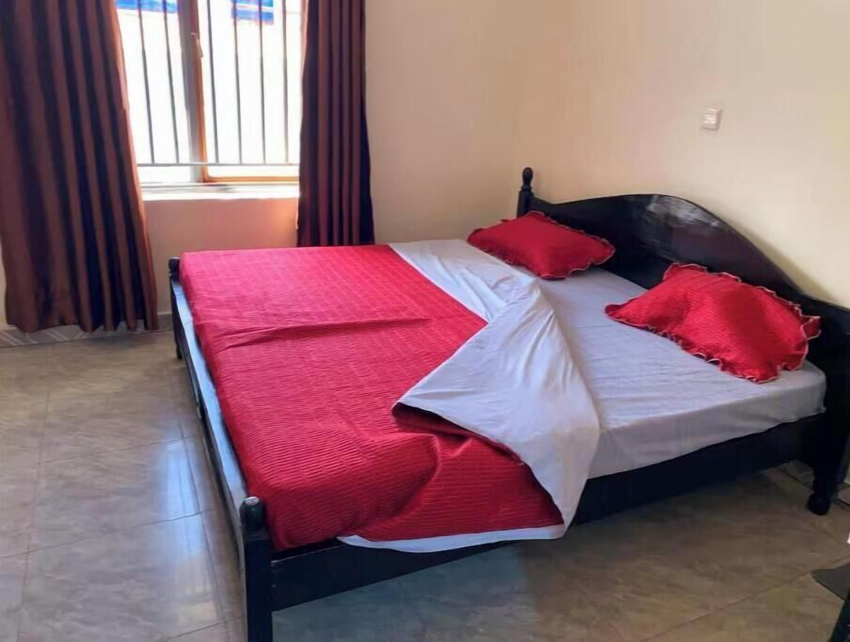 Una cama con estructura negra, ropa de cama roja y blanca, y una ventana con cortinas.